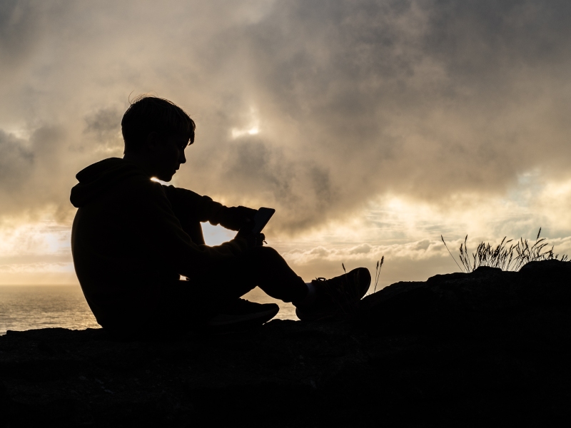 A silhouette of a boy as he sits atop a wall checking his social media by the Atlantic_Ocean
